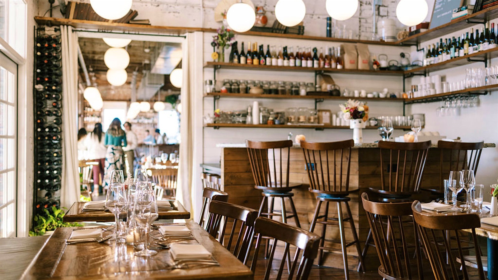 Set tables and a bar setup, made of wood, at Ronin, with white brick walls and wine bottles on shelves and racks along the wall; spherical light fixtures hang overhead, with people shown in another room in the background